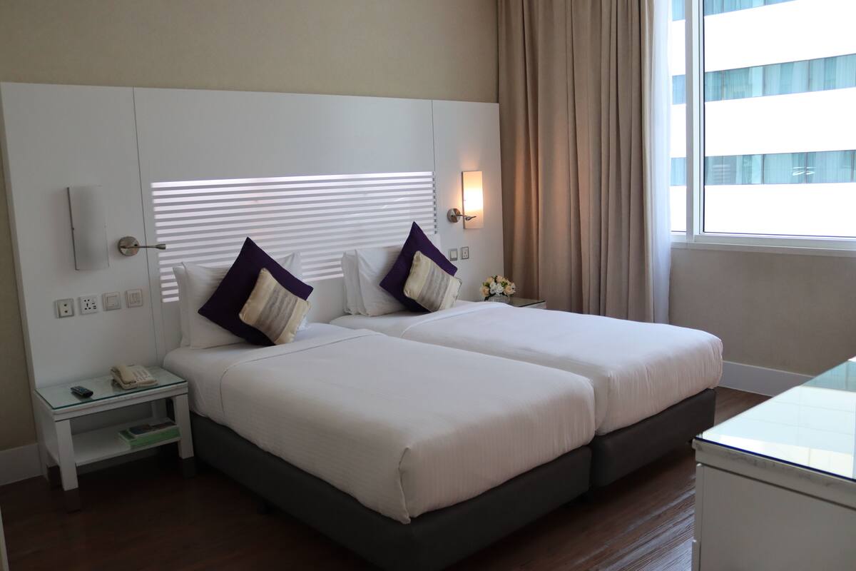 A hotel room with two beds, a bedside table with a phone, a nightstand, and a window with curtains.