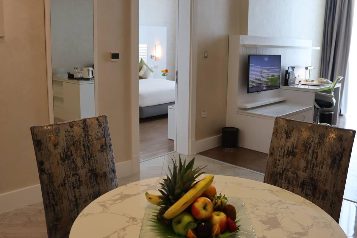A hotel room with a dining table set with fruit, a bed, a TV, and a desk with a computer.