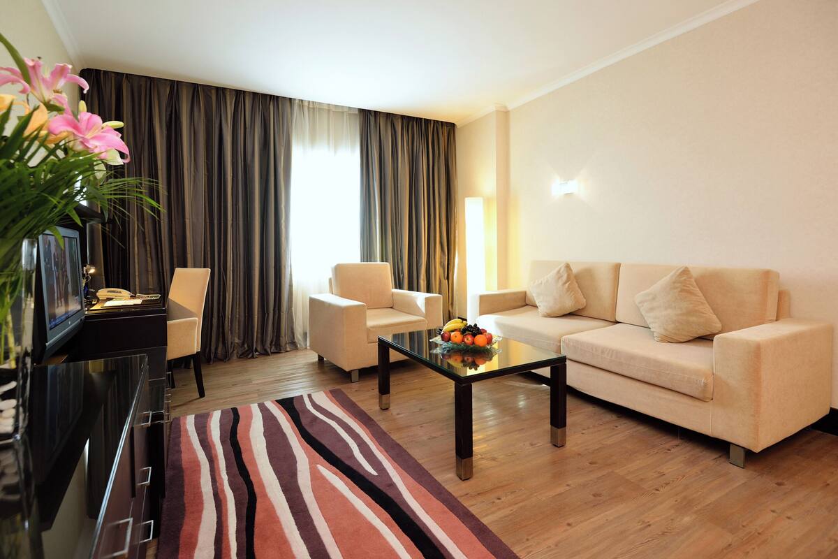 A modern living room with a sofa, armchairs, a coffee table with fruit, a TV, and a large window with curtains.