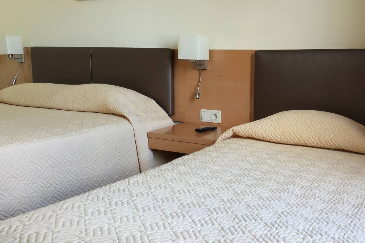 A hotel room with two beds, each with a headboard and a bedside table. There is a remote control on one of the tables.