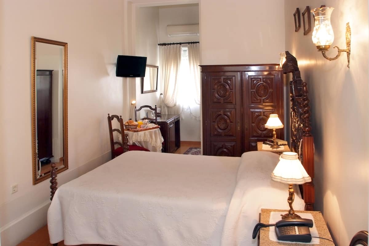 A hotel room with a large bed, a wooden wardrobe, a bedside table with a lamp, a mirror, and a TV mounted on the wall.
