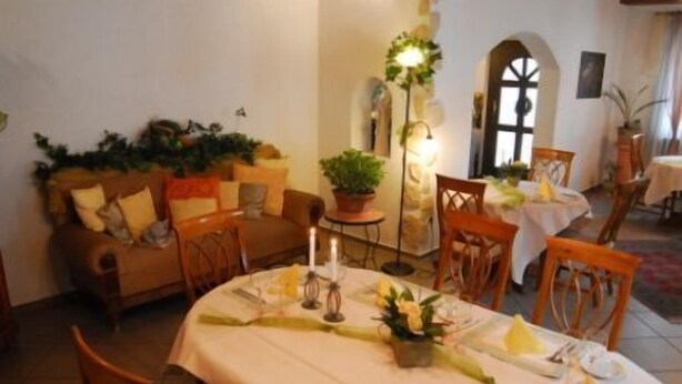 A dining area with a round table set for four, surrounded by wooden chairs. A sofa with cushions is against the wall, and a potted plant is on a side table. - Hotel Restaurant Linde (Otterstadt)