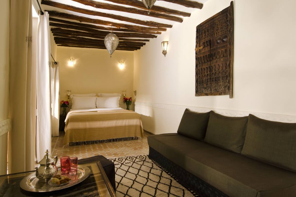A cozy bedroom with a bed, a sofa, a tray with a teapot and glasses, and a decorative wall piece.