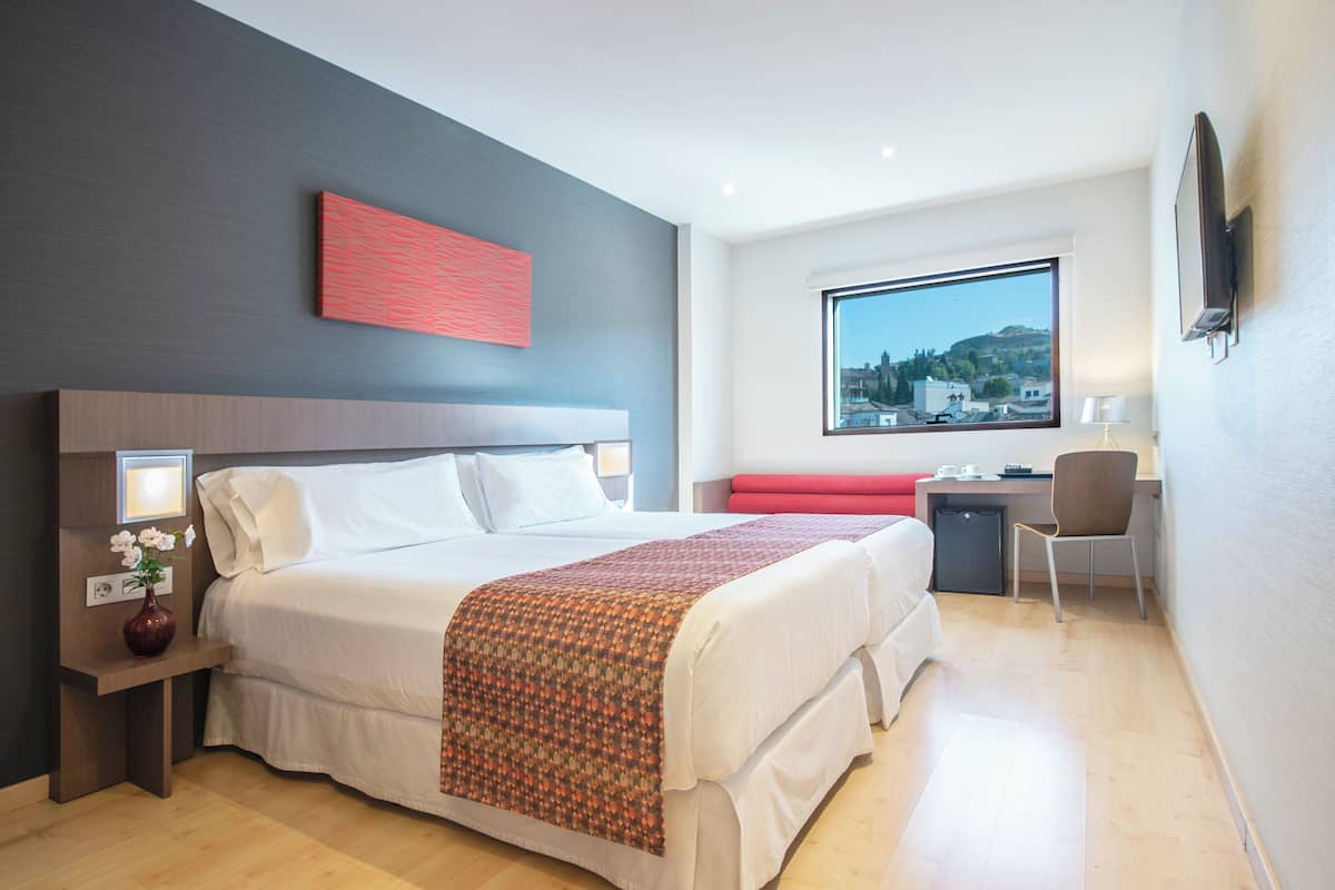 A hotel room with a large bed, a desk, a chair, and a view of a cityscape through a window.