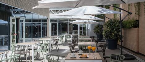 Un café en plein air avec des tables et des chaises, des parasols, et un bâtiment en briques en arrière-plan.
