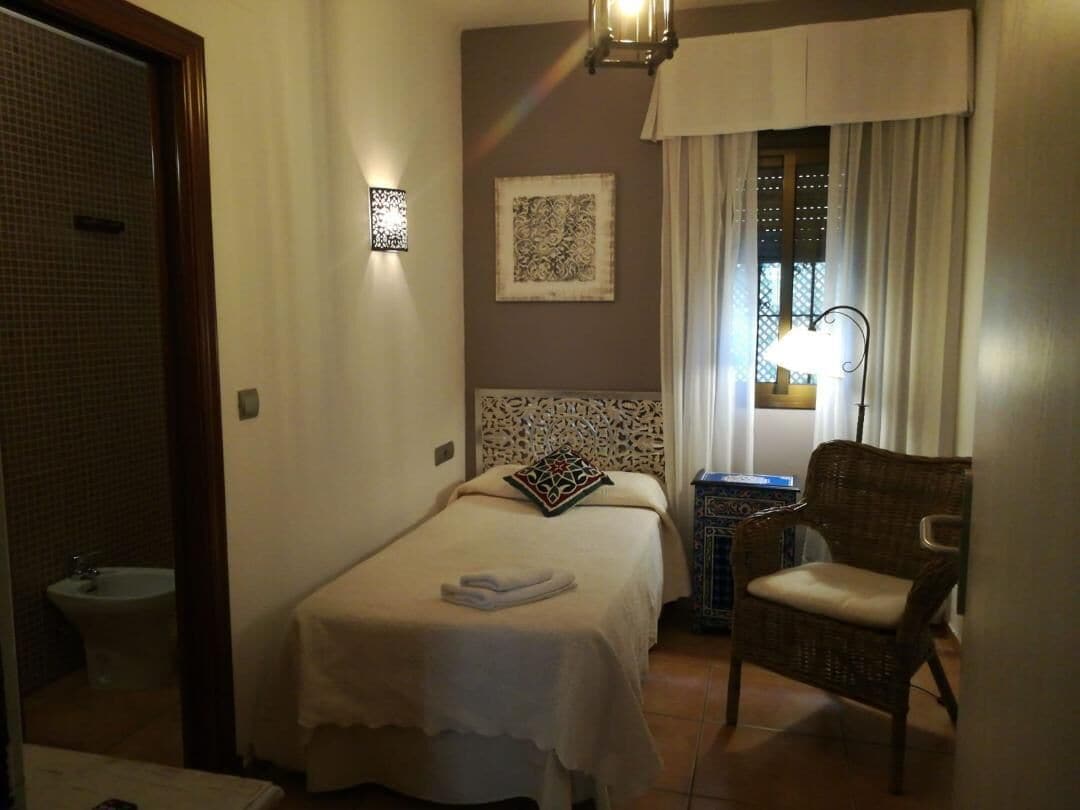 Habitación de hotel con cama, una silla, mesita de noche, lámpara y ventana con cortinas.