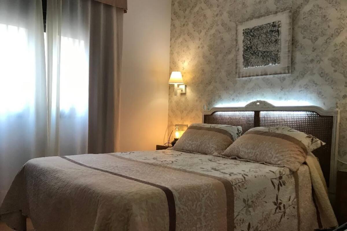 A neatly made bed with a floral bedspread, a wall-mounted lamp, and a framed artwork above the bed.