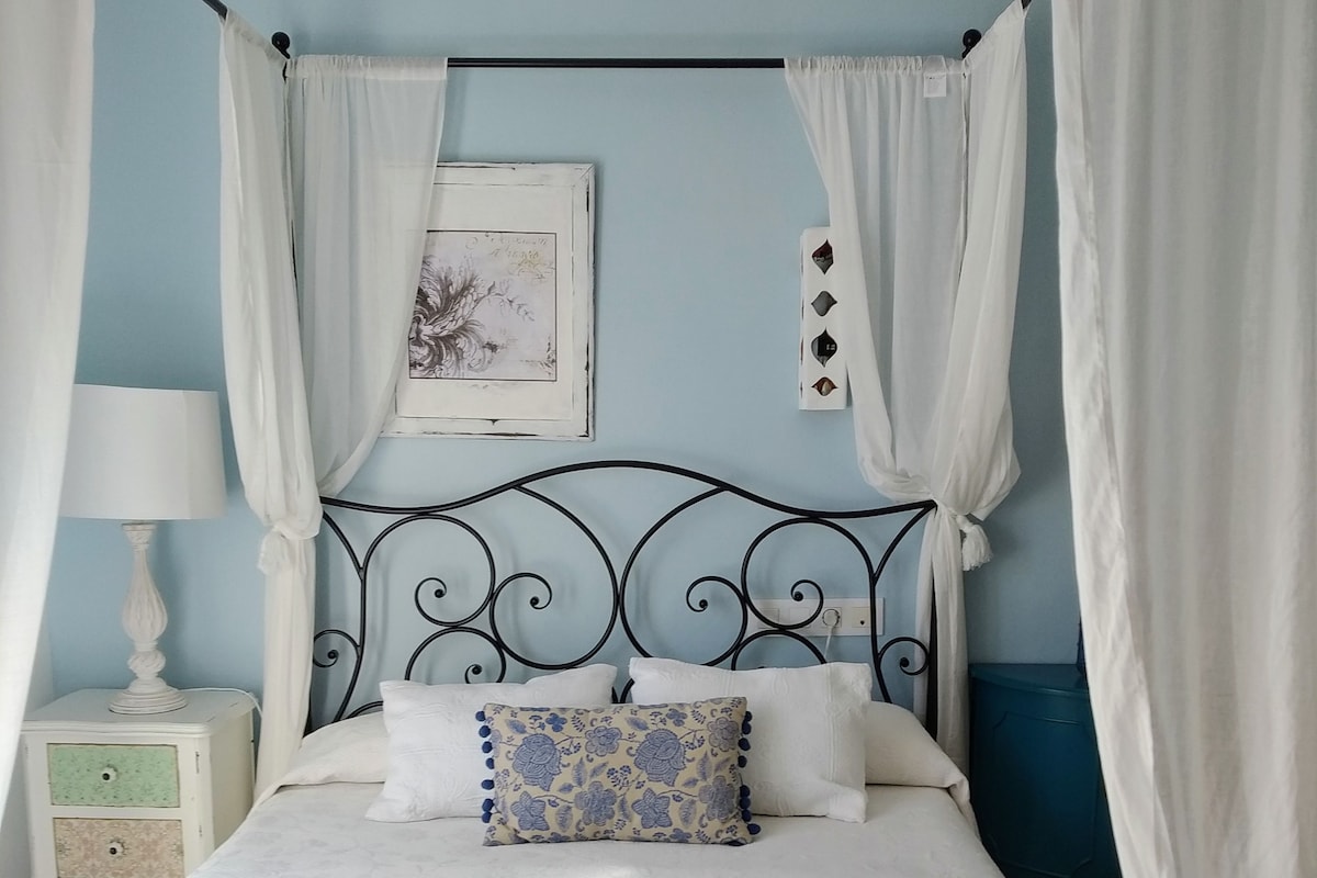 Un lit avec un cadre en métal, une literie blanche et un coussin à motifs floraux. Il y a une table de chevet avec une lampe, et un cadre photo est accroché au mur.