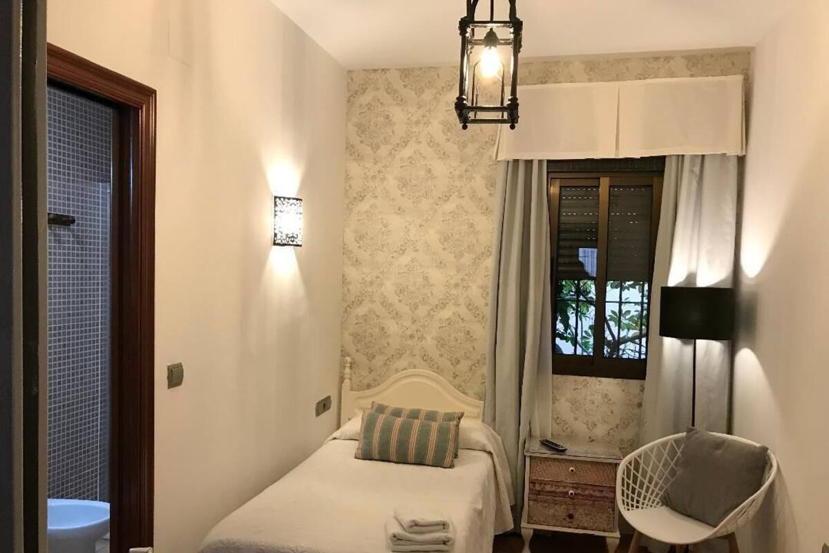 A bedroom with a bed, a chair, a nightstand, a window with curtains, and a wall-mounted light fixture.
