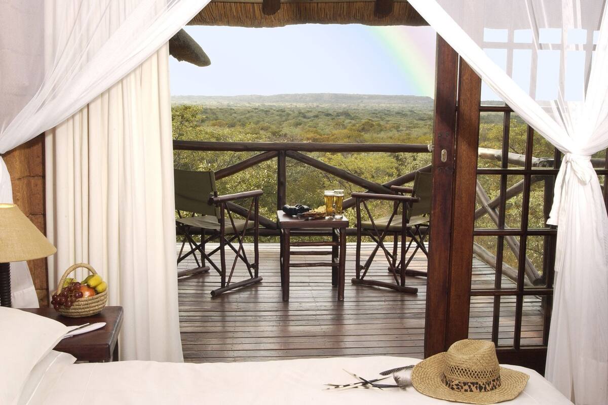 A room with a bed, a table with drinks, a basket of fruit, a straw hat, and a view of a landscape with trees and a rainbow.