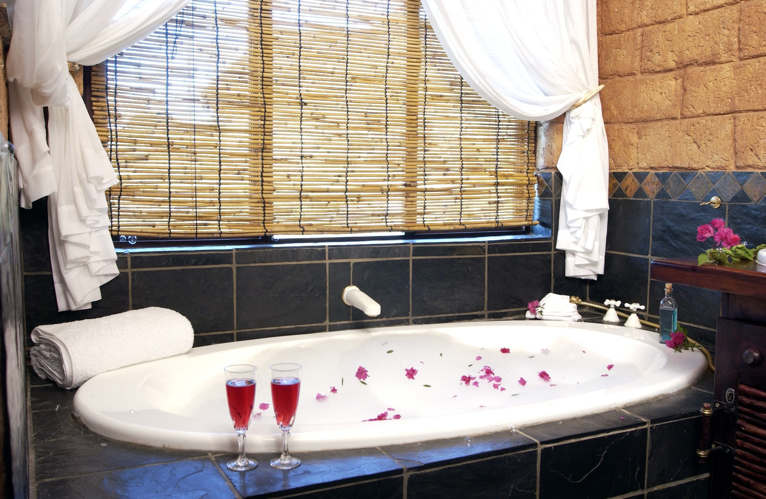 A bathroom with a bathtub, two glasses of red liquid, and a window with bamboo blinds.