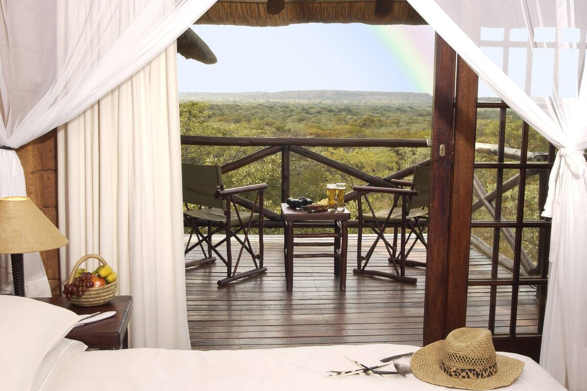 A room with a bed, a bedside table with a lamp and fruit basket, a wooden deck with a table and chairs, and a view of the landscape.