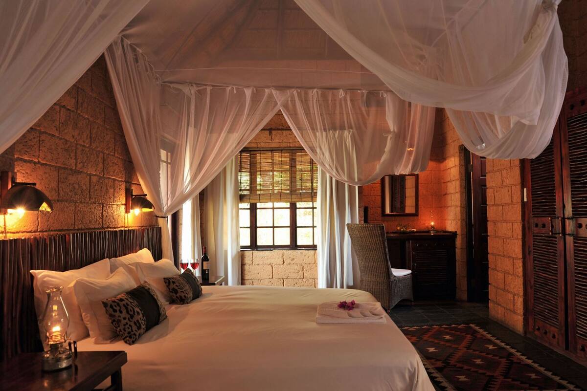 A bedroom with a bed, a nightstand, a lamp, a window, and a mirror.