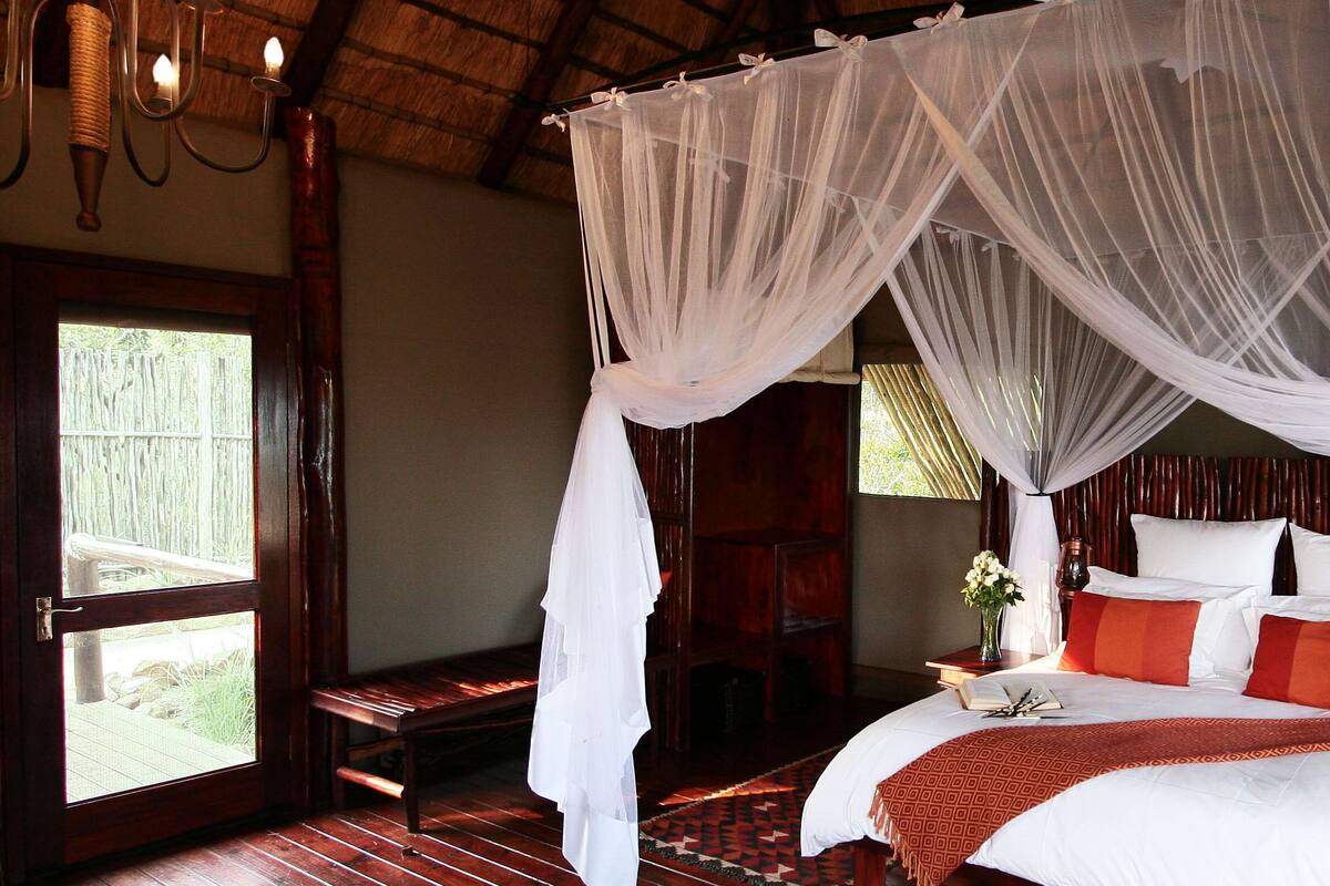 A bedroom with a canopy bed, wooden flooring, a bench, and a view of the outdoors.