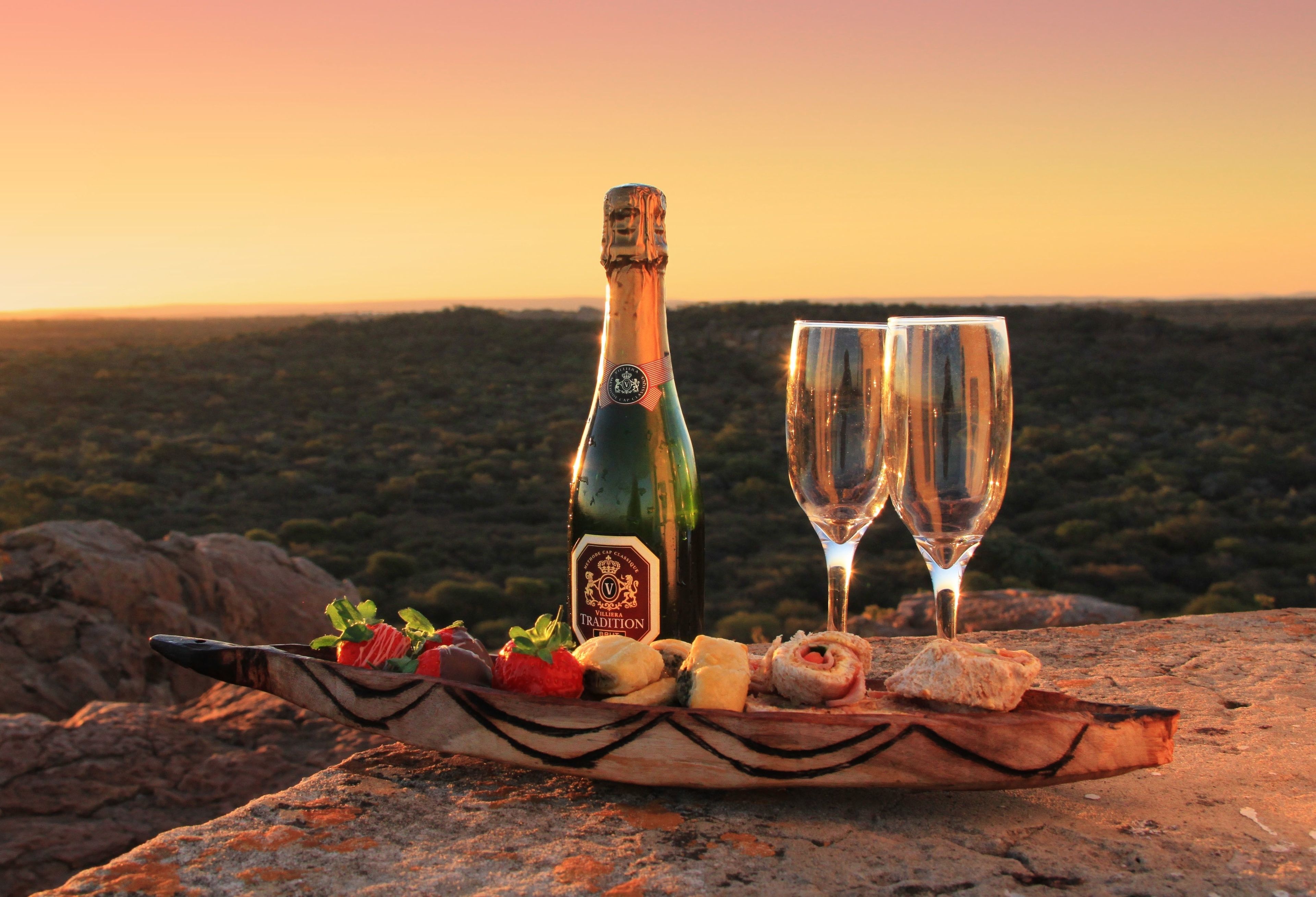A bottle of wine and two glasses on a wooden board with snacks, set against a scenic sunset background.