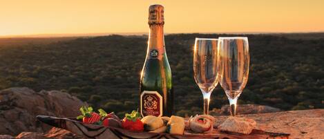 A bottle of wine and two glasses on a wooden board with snacks, set against a scenic sunset background.