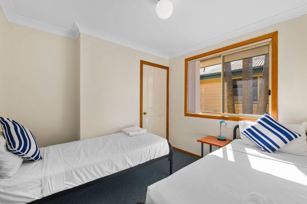 A hotel room with two beds, a small table with a lamp, a window with blinds, and a door.