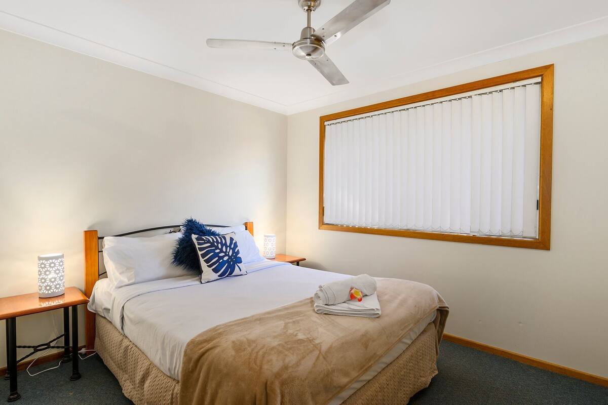 A neatly made bed with a beige blanket, a blue pillow, and a white pillow. There is a ceiling fan above the bed, a bedside table with a lamp, and a window with blinds.