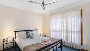 Une chambre à coucher avec un lit, un ventilateur de plafond, une table de chevet avec une lampe et une fenêtre avec des rideaux légers.