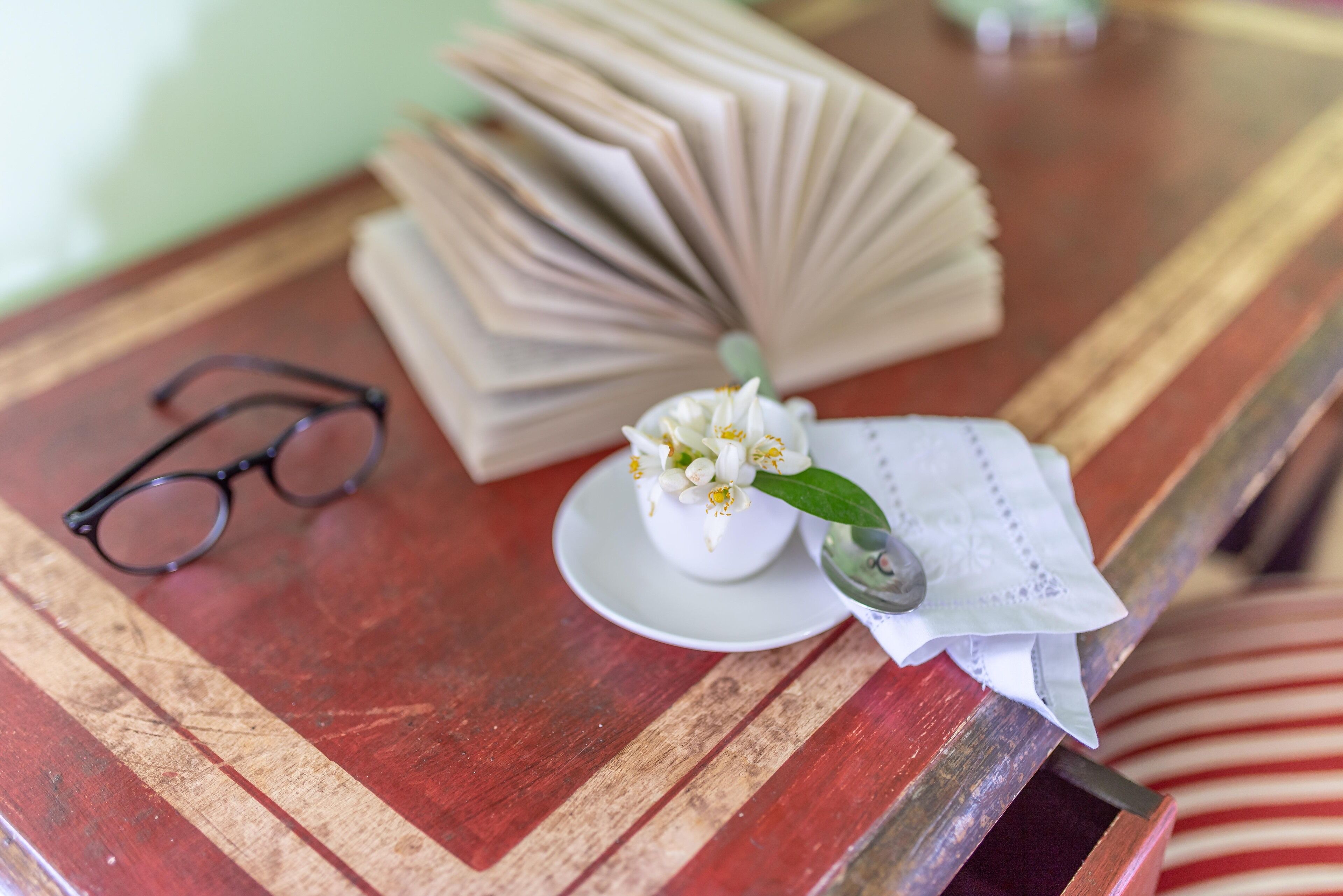 Una mesa de madera con un par de gafas, un libro doblado, una taza blanca pequeña con una flor y una cuchara.