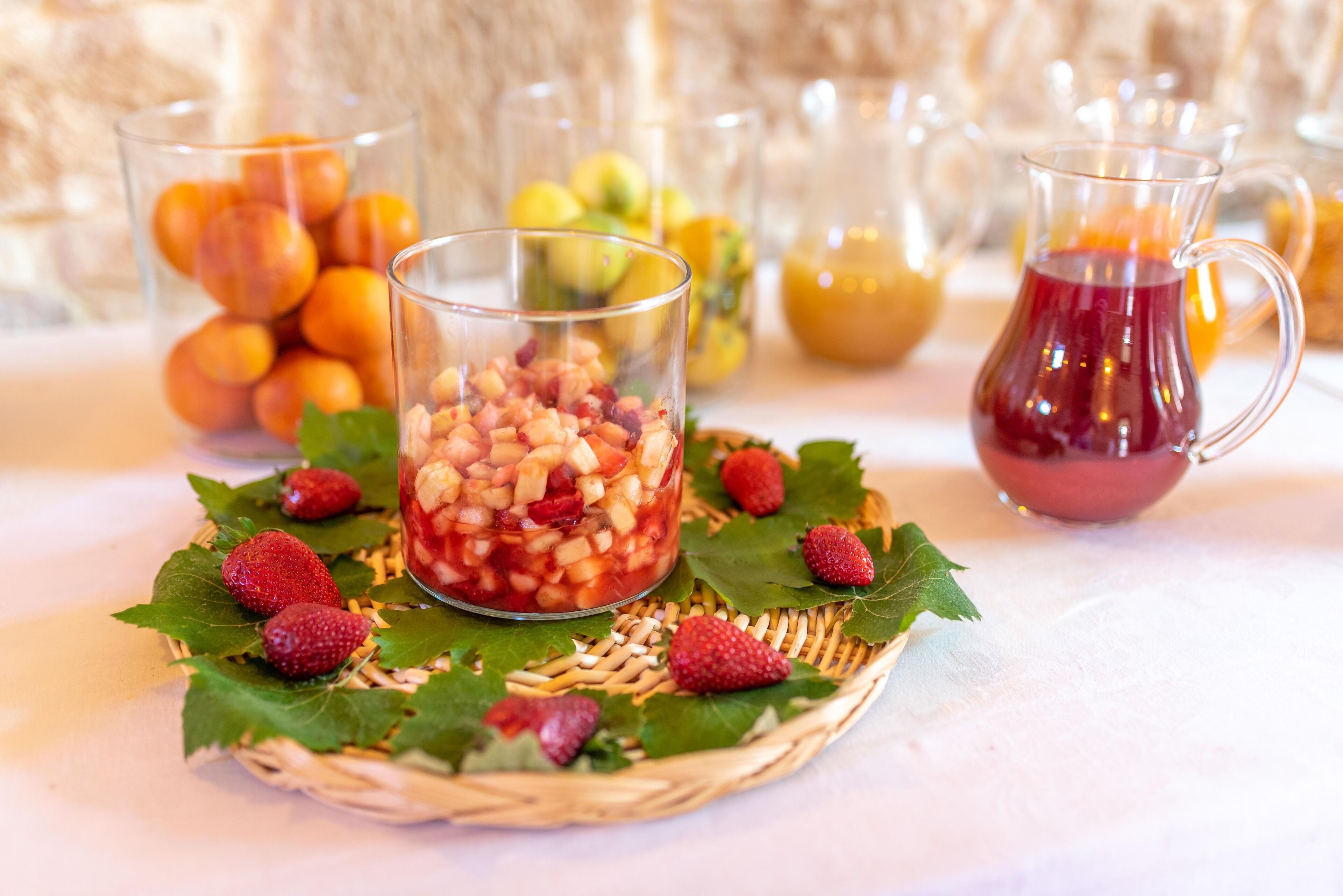Ein Tisch mit einem Glasbehälter voller gewürfelter Früchte, umgeben von Erdbeeren und Blättern, mit verschiedenen Früchten und einer Kanne roter Flüssigkeit im Hintergrund.