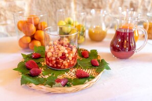 Un tavolo con un recipiente di vetro pieno di frutta a cubetti, circondato da fragole e foglie, con altra frutta e una brocca di liquido rosso sullo sfondo.