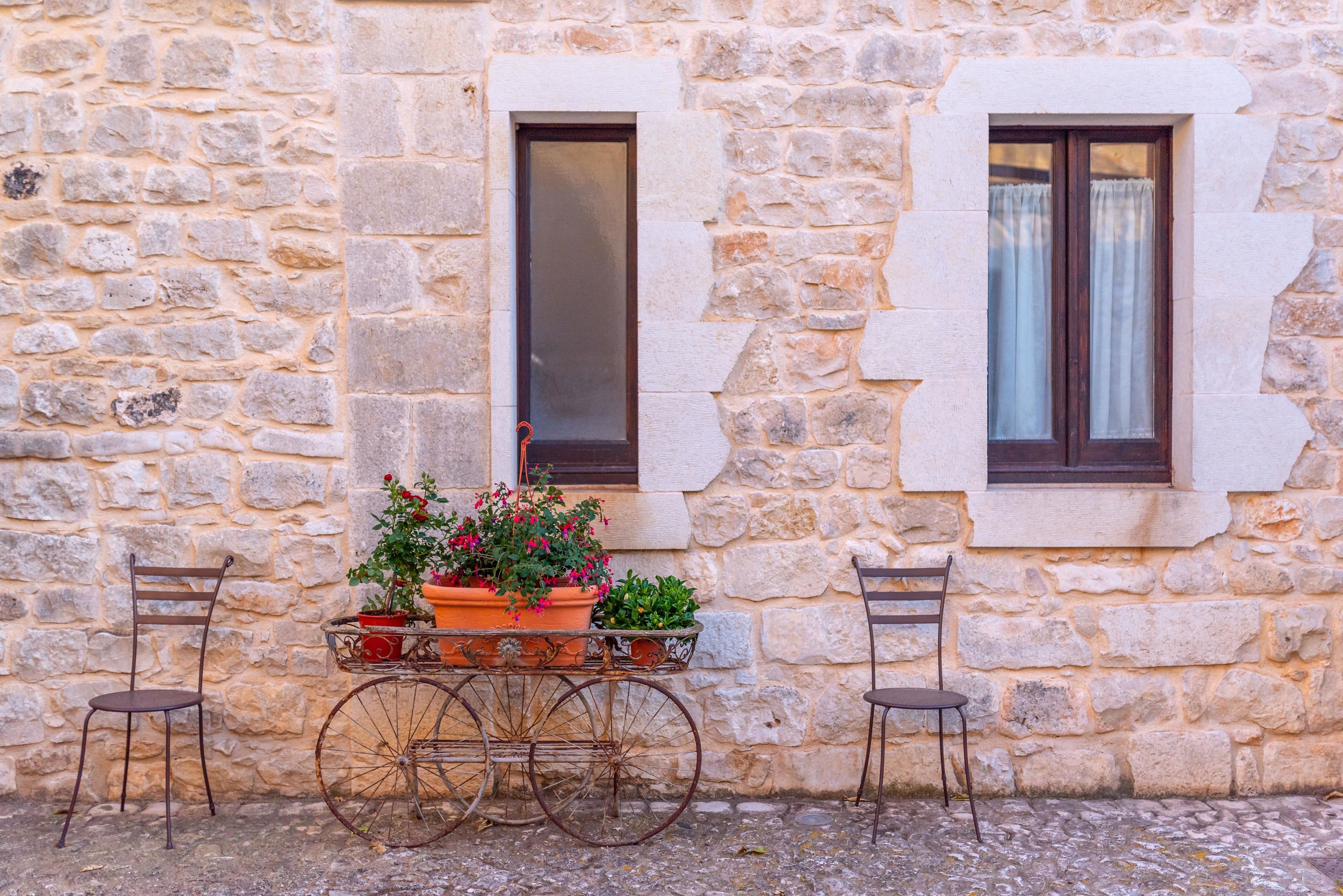 A stone building with two windows, a cart with potted plants, and two chairs.