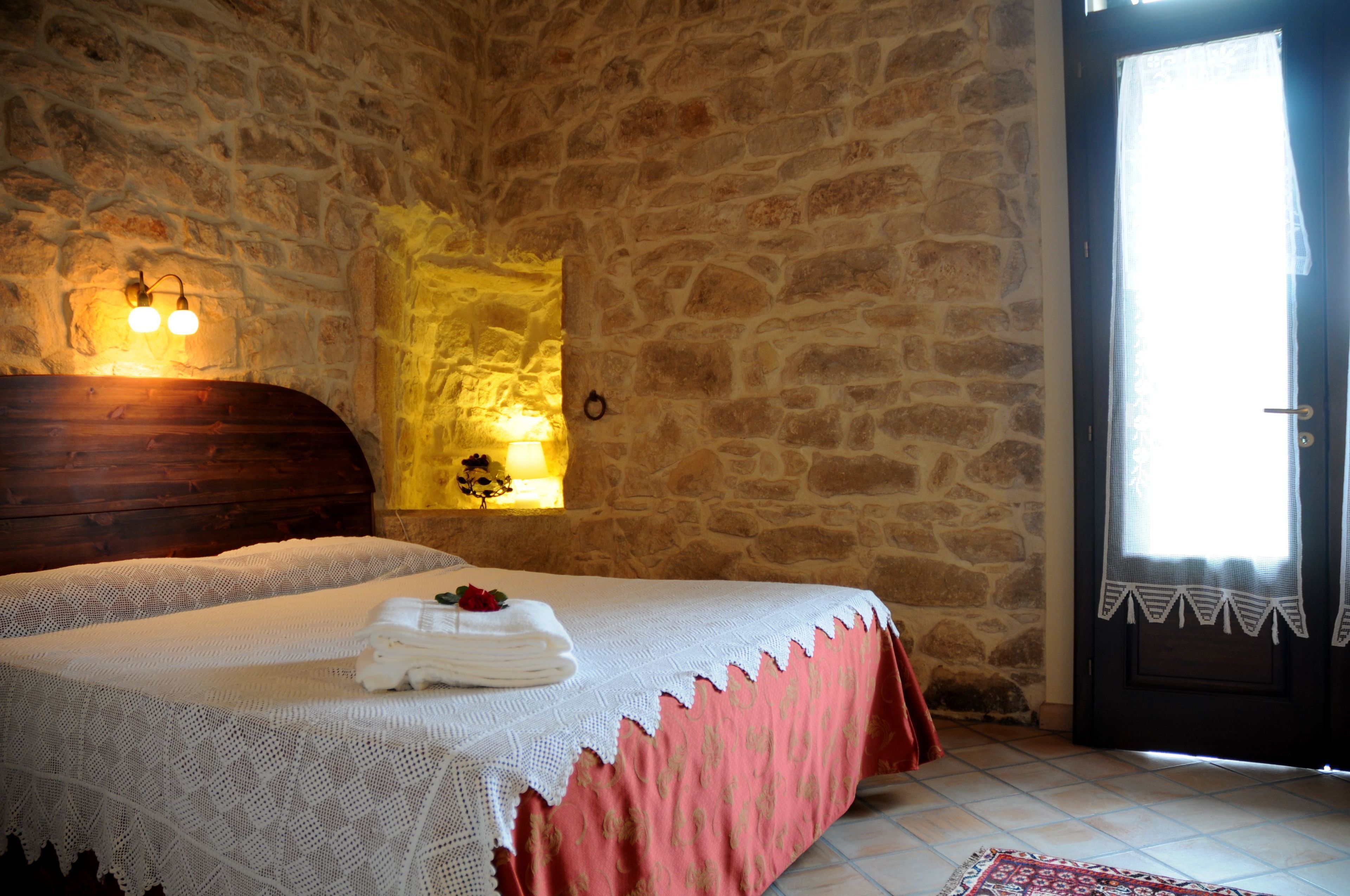 Une chambre avec un mur en pierre, une tête de lit en bois, un lit recouvert d’une couvre-lit blanche et rose, et une table de chevet avec une lampe.