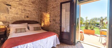 A bedroom with a stone wall, a bed with a red blanket, and a view of the garden through an open door.