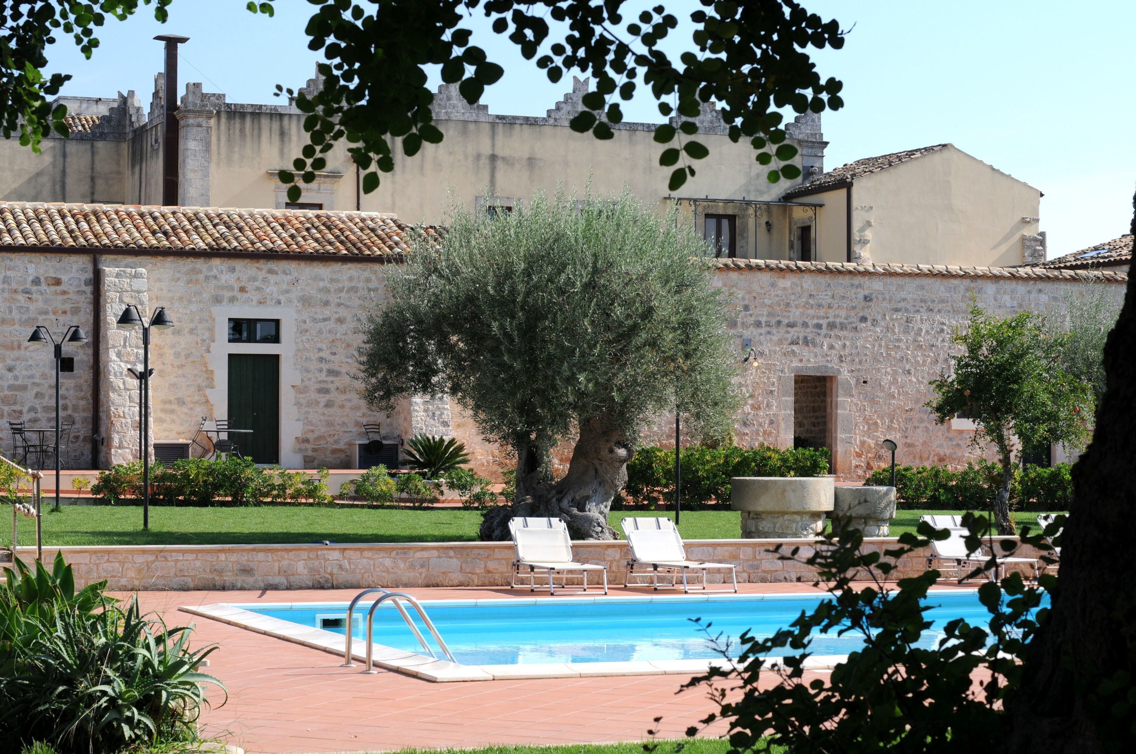 Un área de piscina con sillones, un edificio de piedra con tejado de baldosas y un árbol grande.