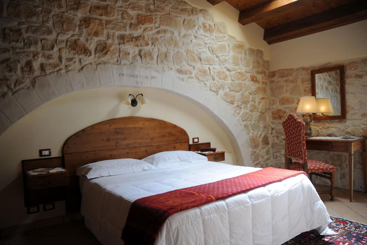 A bedroom with a stone wall, wooden bed frame, a desk with a chair, and a lamp.