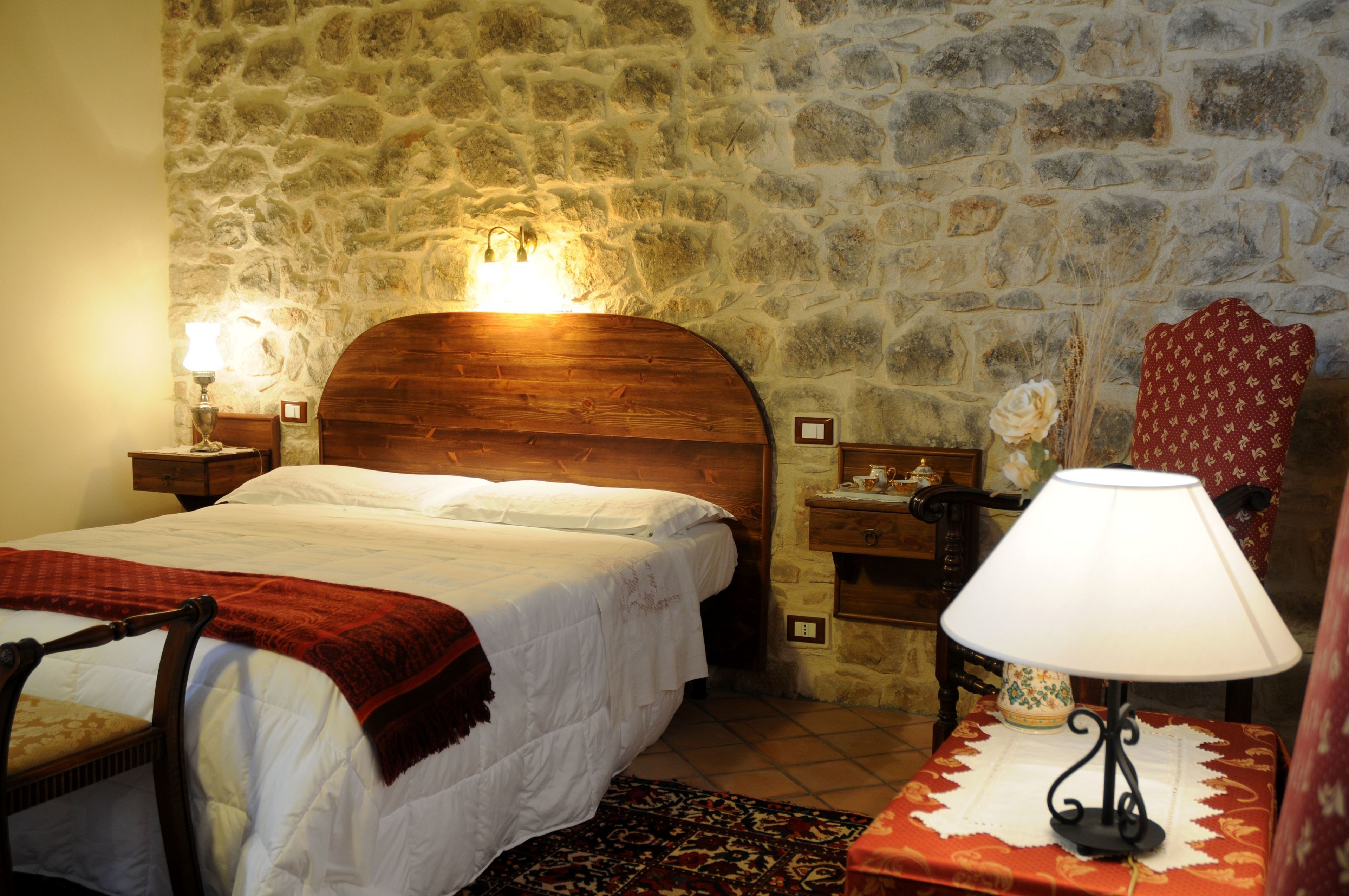 Un dormitorio con pared de piedra, cabecero de madera, cama con ropa de cama blanca, una manta roja, una mesita de noche con una lámpara y una pequeña mesa con un jarrón de flores.