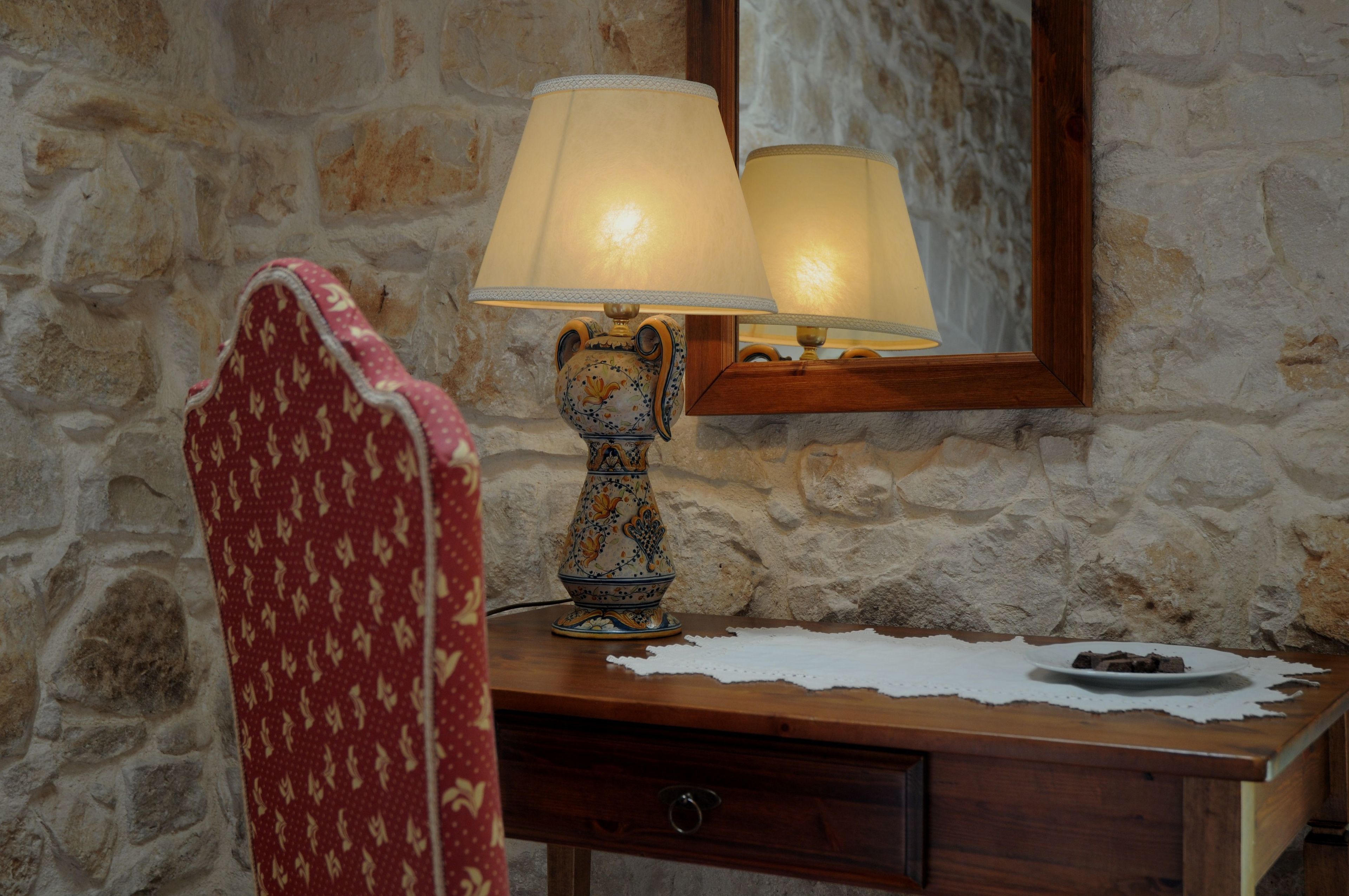 A wooden desk with a lamp, a mirror, and a chair with a red patterned cushion.