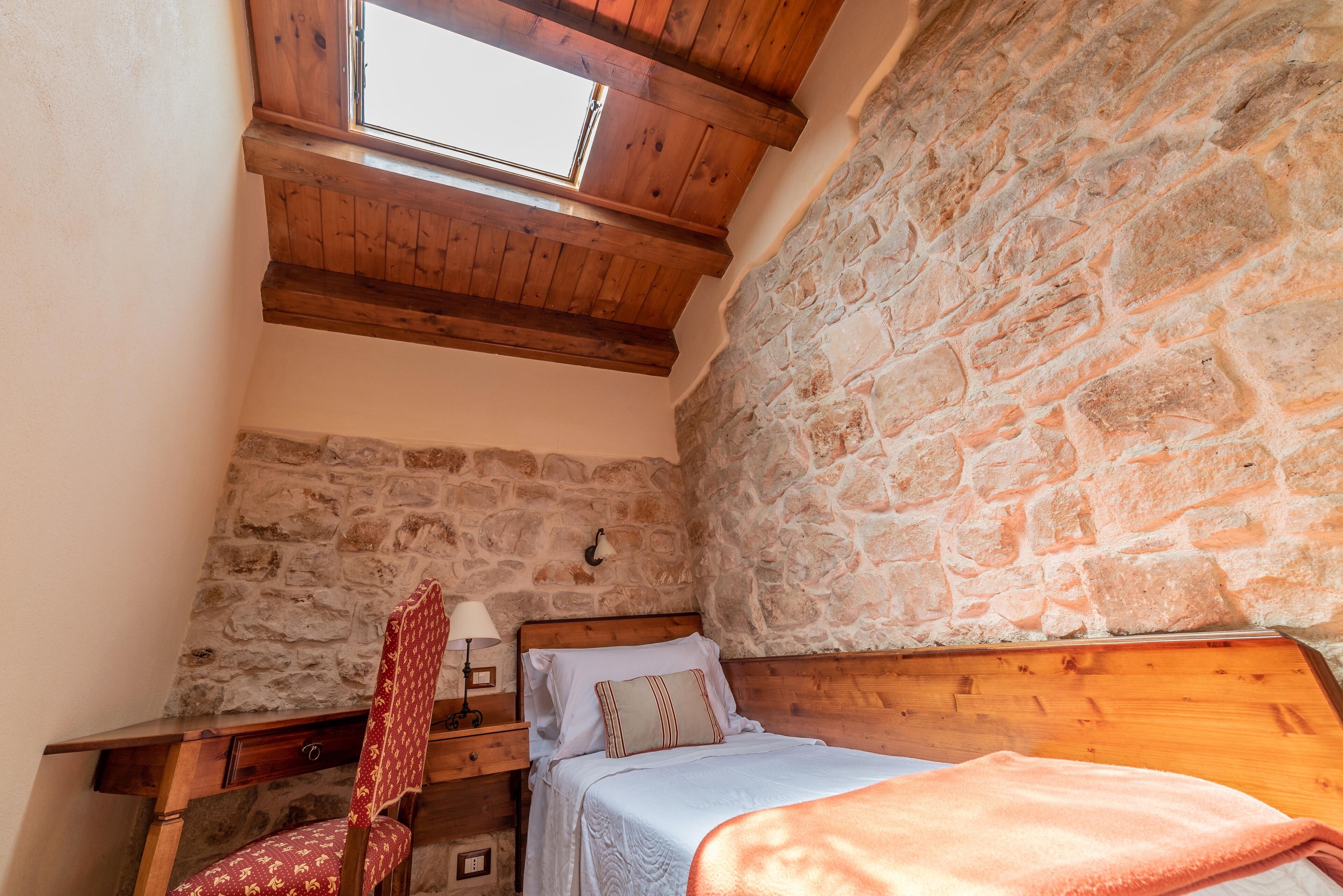 Une chambre avec un mur en pierre, des poutres en bois au plafond, un lit, une chaise et un bureau.