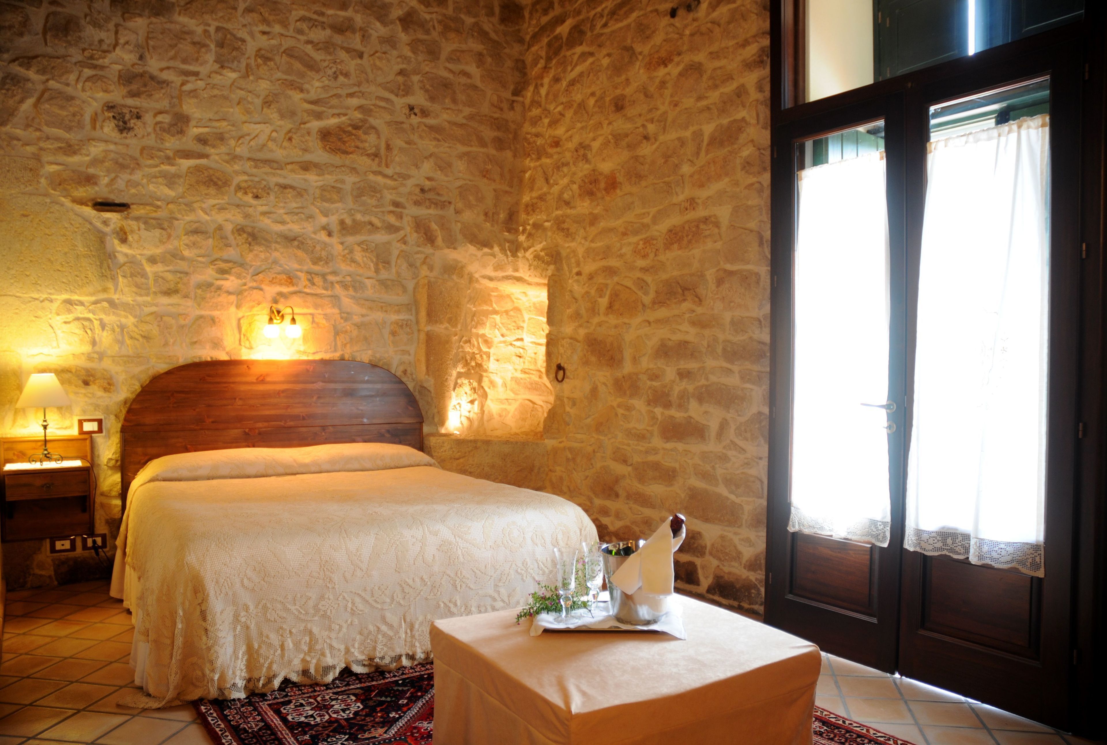 A bedroom with a stone wall, a wooden headboard, a bed with a white bedspread, a nightstand with a lamp, and a window with curtains.