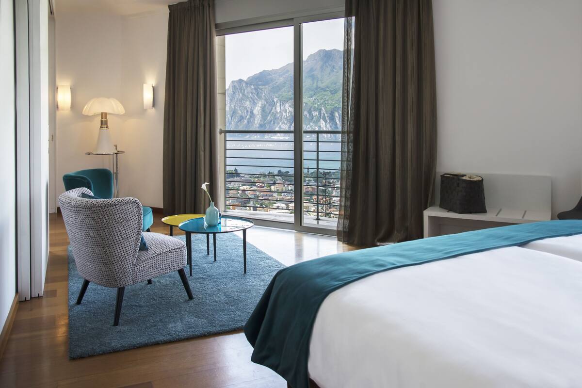 A hotel room with a large bed, a chair, a small table, and a view of the sea and mountains through the window.