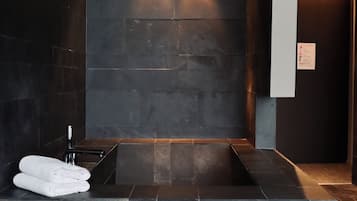 Bathroom features a large bathtub and dark tile walls with a towel rack and folded towels.