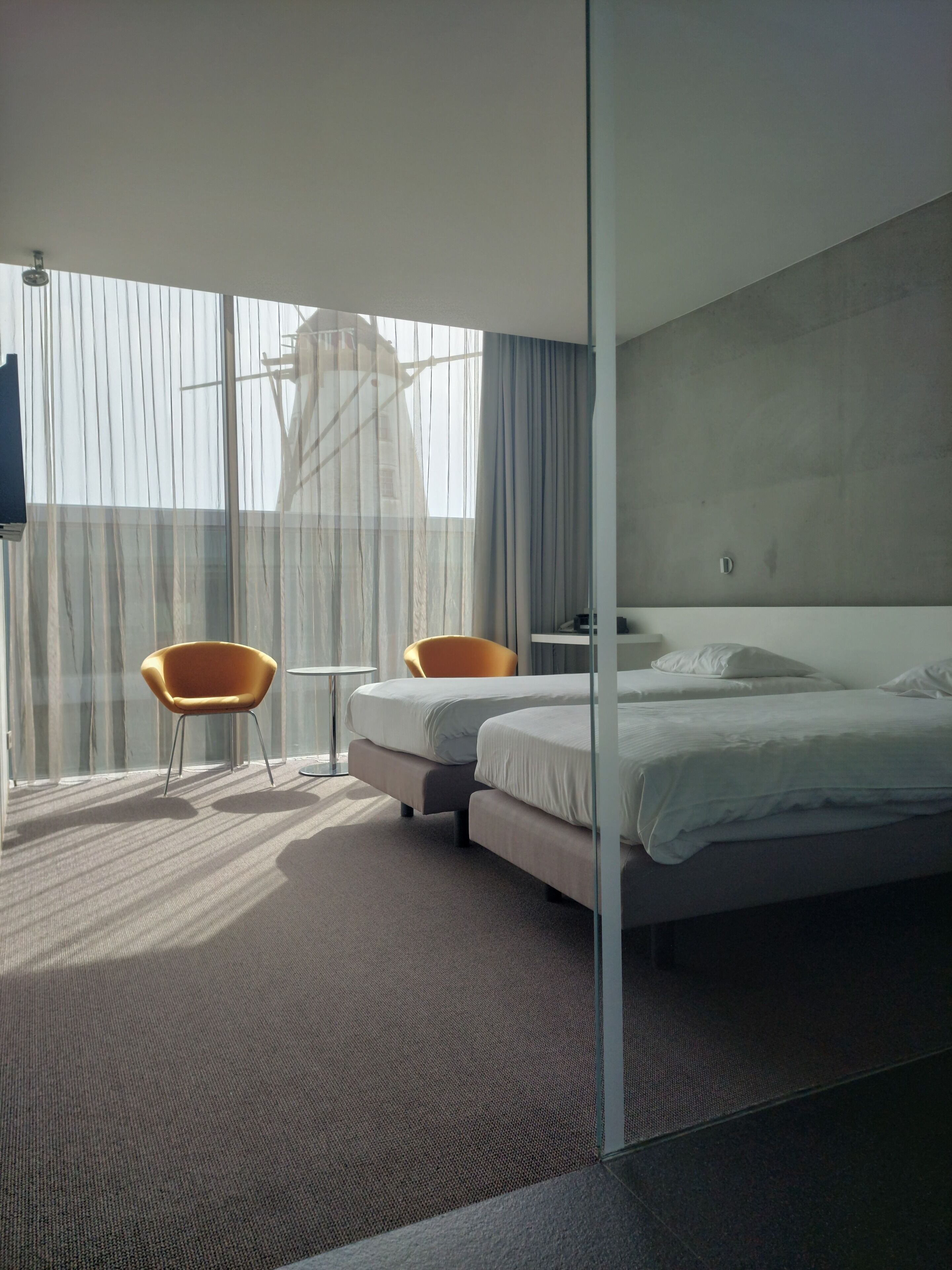 A modern hotel room with a large bed, two chairs, a small table, and a large window with curtains.