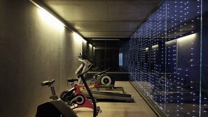 Une salle de sport équipée de vélos d’appartement et de tapis de course, avec un plafond en béton et un mur orné d’une grille de lumières.