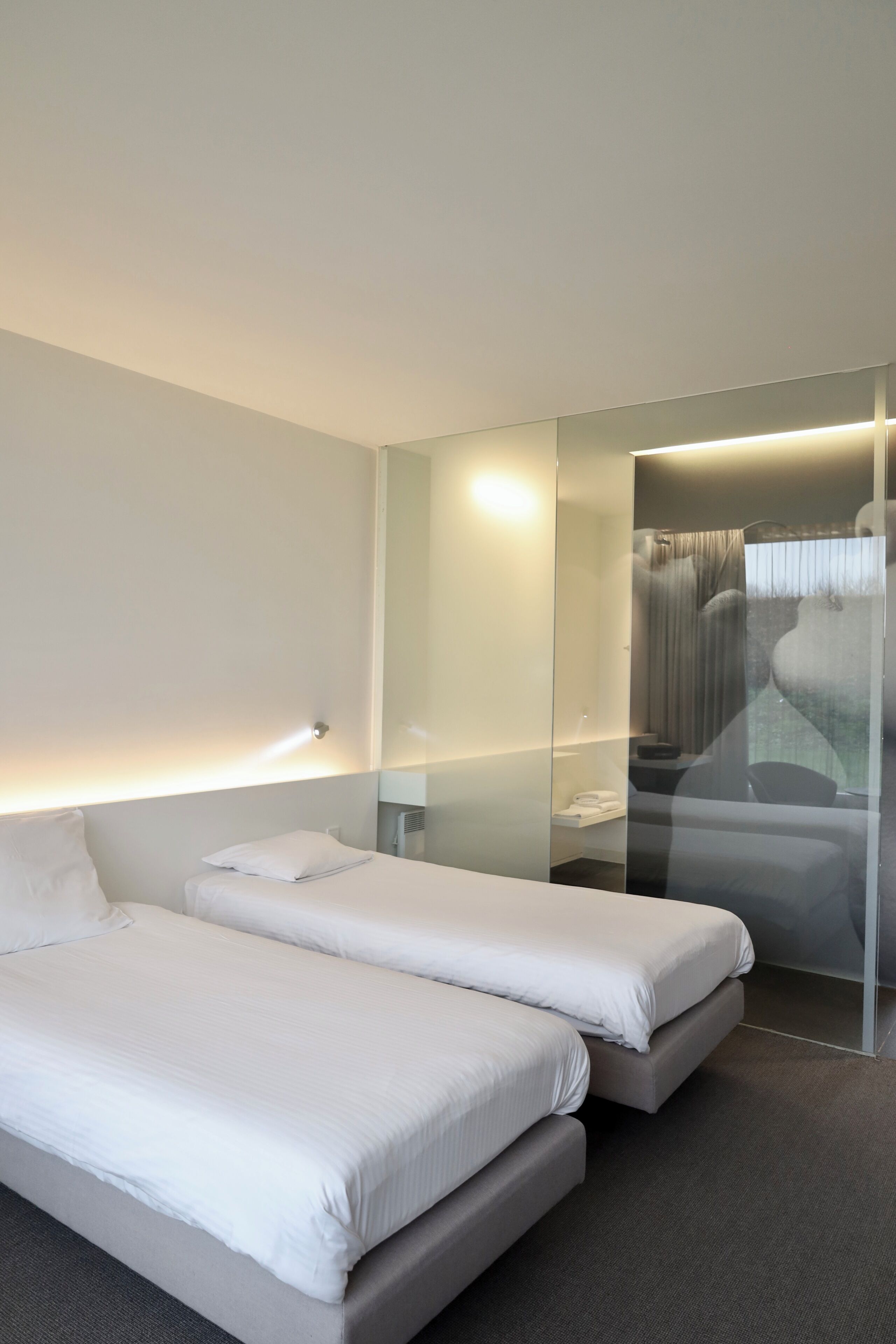 Une chambre d’hôtel moderne avec deux lits, une cloison en verre et une salle de bain visible.