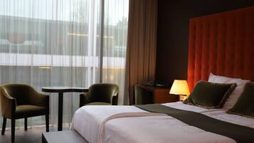 Ein Hotelzimmer mit einem großen Bett, zwei Sesseln, einem kleinen Tisch und Blick auf die Stadt durch große Fenster.