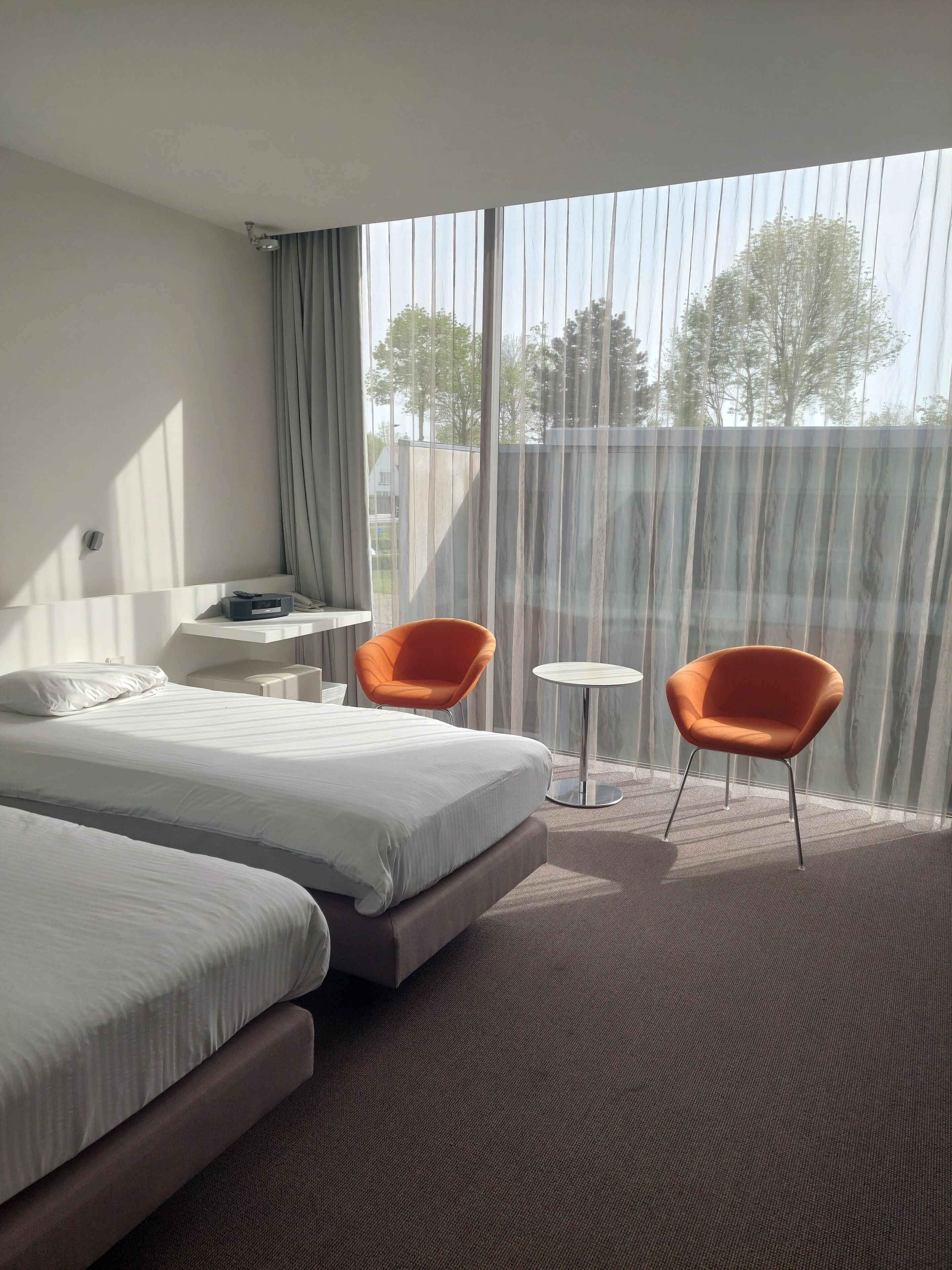 A hotel room with two beds, an orange chair, a small table, and a large window with curtains.