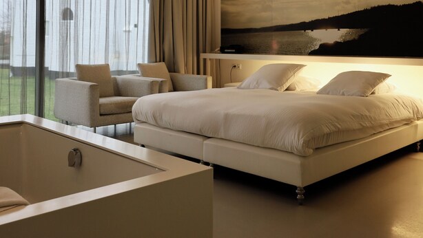 Une chambre moderne avec un grand lit, un coin salon avec des fauteuils, une baignoire et une vue magnifique sur un lac, visible par la fenêtre.