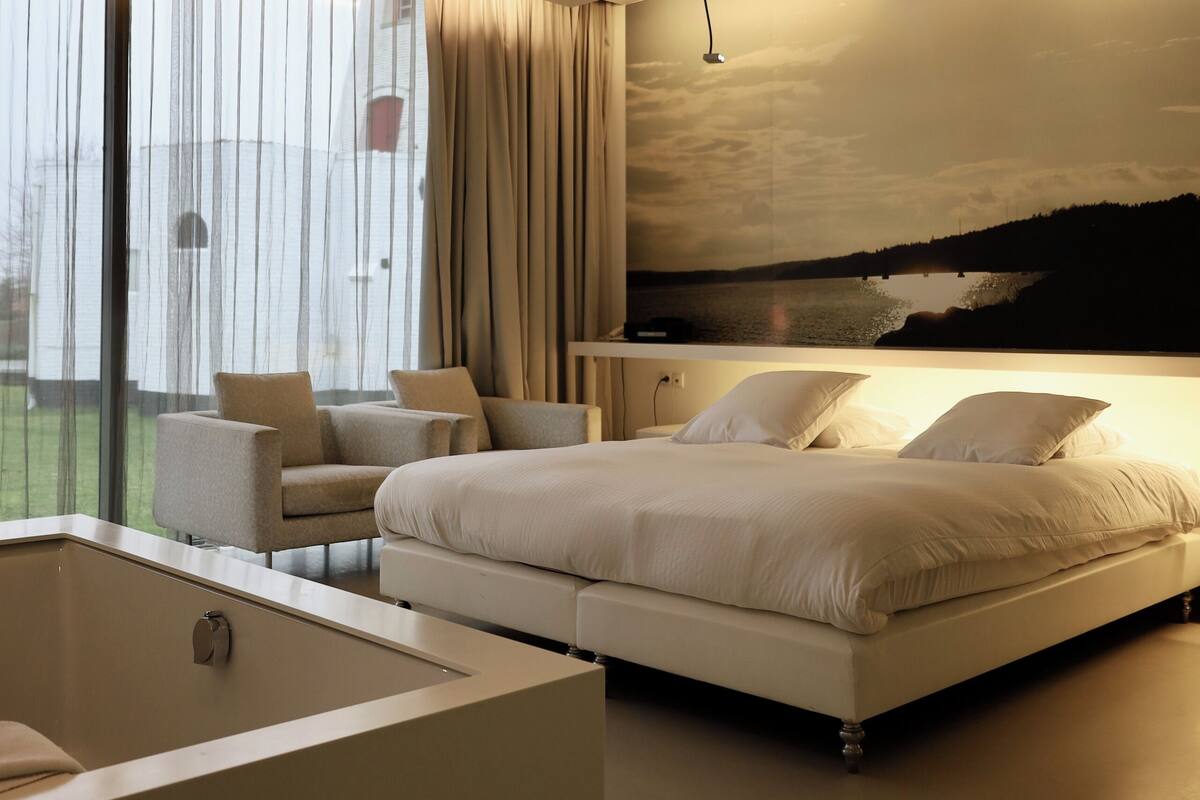 A modern bedroom with a large bed, a sitting area with chairs, a bathtub, and a scenic view of a lake through the window.