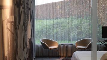 A hotel room with a large bed, two chairs, a small table, and a view of a forest through sheer curtains.