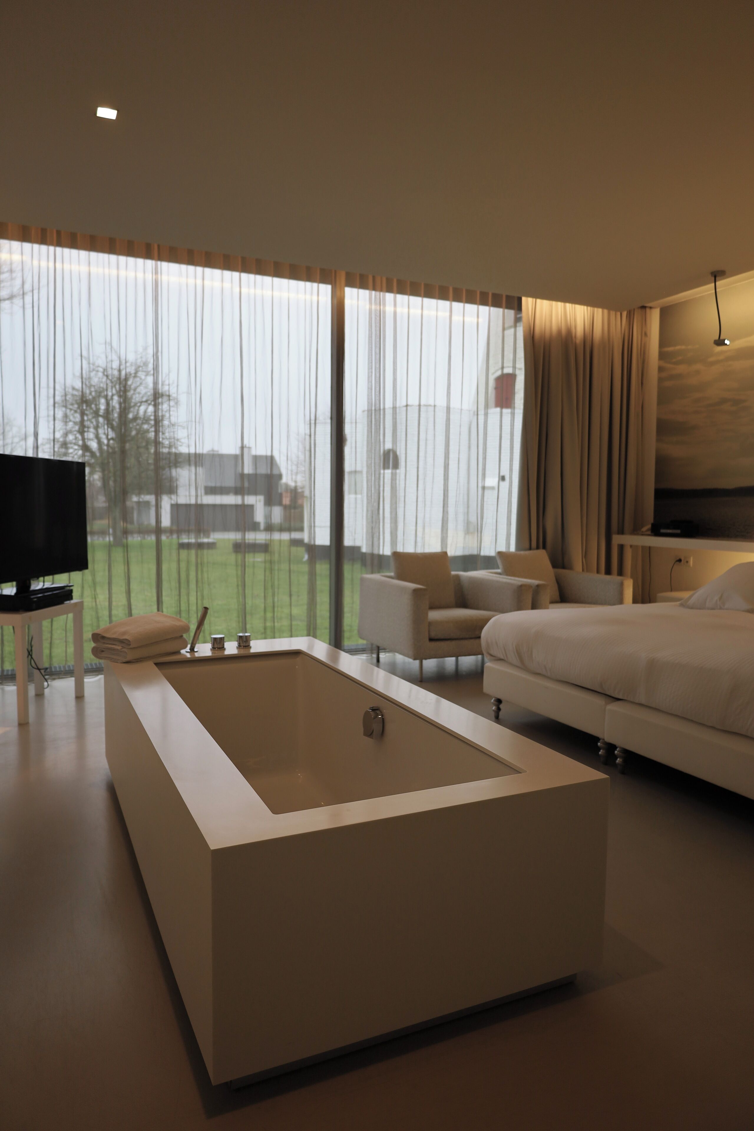 A modern hotel room with a large window, a flat-screen TV, a sofa, a bed, and a bathtub.