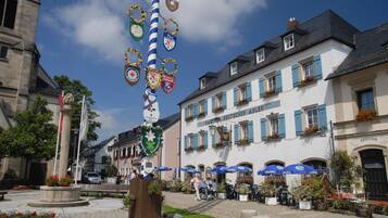 Una plaza tradicional de pueblo europeo con una fuente central, rodeada de edificios históricos con contraventanas azules y jardineras llenas de flores.