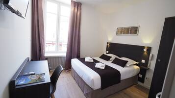 A hotel room with a bed, bedside table, desk, chair, and a window with curtains.