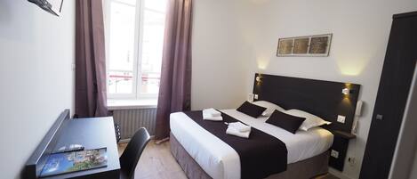 A hotel room with a bed, bedside table, desk, chair, and a window with curtains.