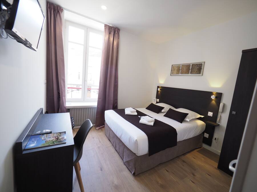 A hotel room with a bed, bedside table, desk, chair, and a window with curtains.